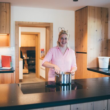 Fröhliche Frau in rosa Bluse lächelt in gemütlicher Holzküche, während sie Kochtopf auf Kochfeld abstellt. Helle Möbel und großes Fenster schaffen freundliche, heimelige Atmosphäre. | © Urlaub am Bauernhof / Daniel Gollner