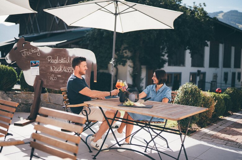 Zwei Menschen sitzen an einem Tisch auf einer Terrasse, ein Sonnenschirm ist aufgespannt, im Hintergrund sind Gartenanlagen zu sehen. Auf dem Tisch stehen Getränke und ein Snack. | © Urlaub am Bauernhof / Daniel Gollner