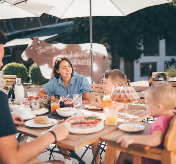Eine Familie mit Kindern genießt ein gemeinsames Mittagessen auf einer Terrasse mit Sonnenschirmen, umgeben von Pflanzen und Dekorationen. | © Urlaub am Bauernhof / Daniel Gollner