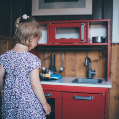 Kleines Mädchen mit blumengemustertem Kleid steht in einer roten, vintage Kinderküche mit Holzwand und modernem Bild an der Wand. | © Urlaub am Bauernhof / Daniel Gollner