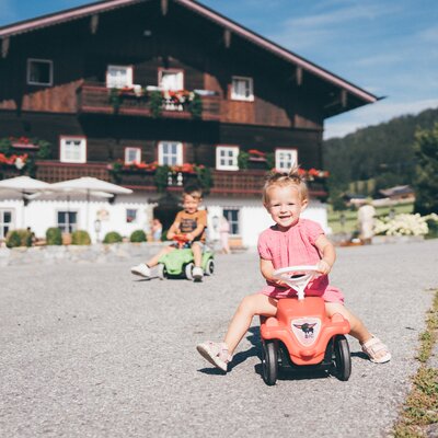 Hier sieht man zwei kleine Kinder, die auf Spielzeugautos auf einer gepflasterten Fläche vor einem großen, traditionellen Gebäude mit Balkonen spielen. Die Umgebung wirkt ländlich und idyllisch. | © Urlaub am Bauernhof / Daniel Gollner