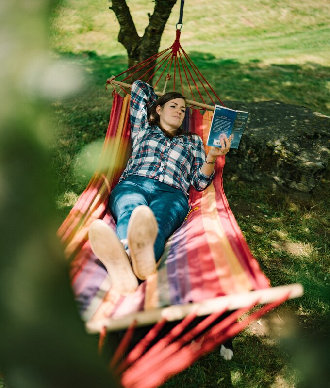 Eine Person, die in einer bunten Hängematte liegend ein Buch liest, umgeben von grüner, üppiger Natur. | © Urlaub am Bauernhof / Daniel Gollner 