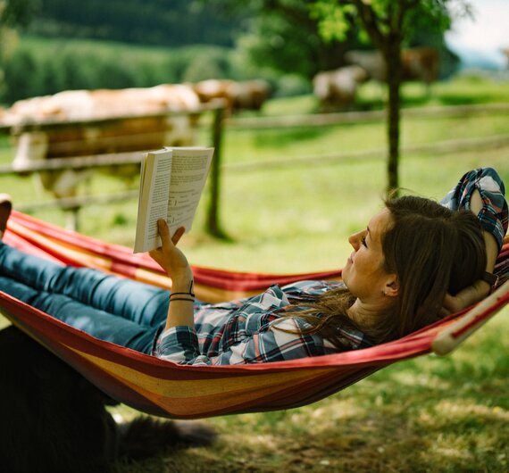 Eine Person entspannt in einer bunten Hängematte in einer grünen Landschaft mit Bäumen und Wiesen, liest konzentriert ein Buch. | © Urlaub am Bauernhof / Daniel Gollner 