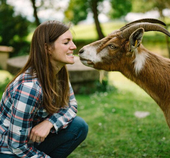 Eine lächelnde Frau in einem karierten Hemd interagiert liebevoll mit einem braunen Ziegenbock in einer idyllischen Umgebung voller Bäume und Wiesen. | © Urlaub am Bauernhof / Daniel Gollner 