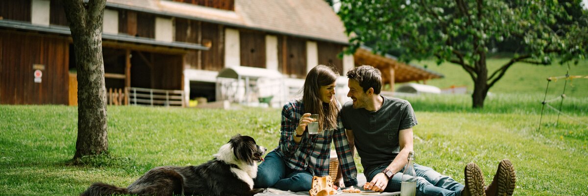Ein junges Paar verbringt einen entspannten Nachmittag auf einer Wiese mit ihrem Hund. Im Hintergrund ist ein rustikales Holzgebäude zu sehen, umgeben von grünen Bäumen. | © Urlaub am Bauernhof / Daniel Gollner 