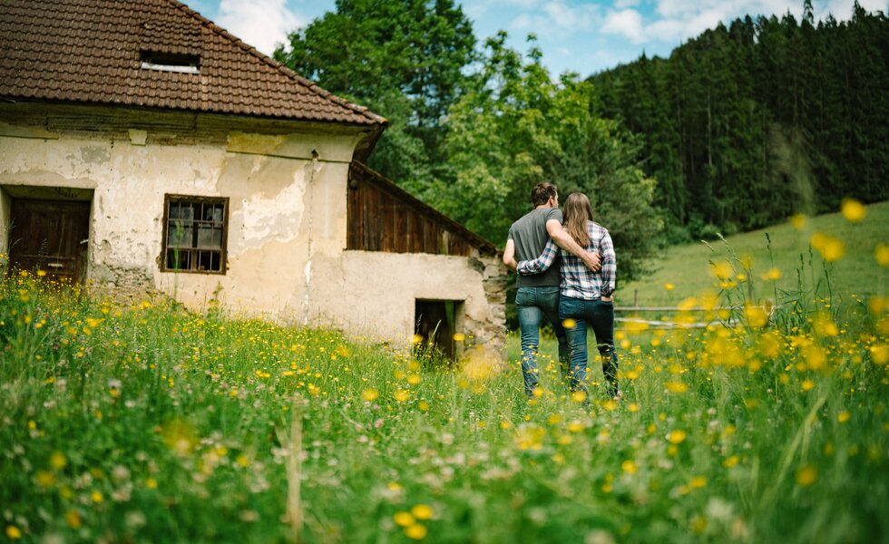 Ein altes Bauernhaus umgeben von einer blühenden Wiese mit Paaren, die arm in arm durch die Natur schlendern. | © Urlaub am Bauernhof / Daniel Gollner 
