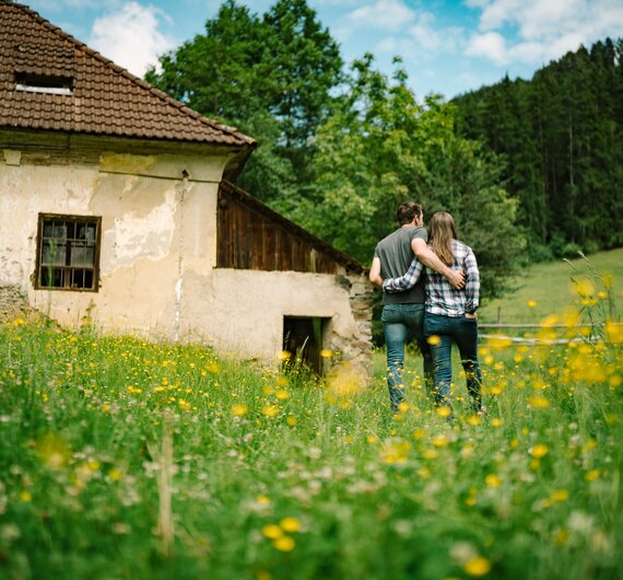 Ein altes Bauernhaus umgeben von einer blühenden Wiese mit Paaren, die arm in arm durch die Natur schlendern. | © Urlaub am Bauernhof / Daniel Gollner 