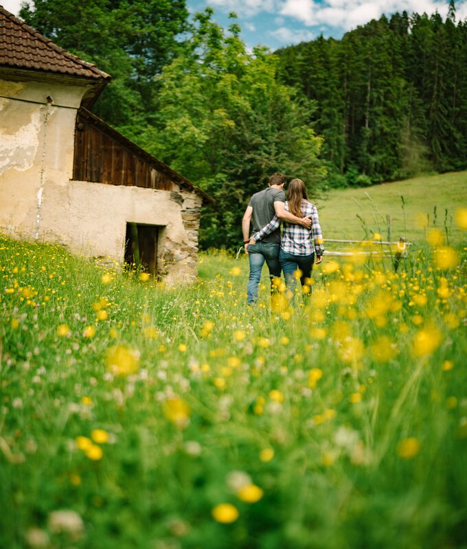 Zwei Personen gehen über eine Wiese voller gelber Blumen, die eine felsige Almhütte umgeben. Im Hintergrund erstreckt sich eine bewaldete Landschaft unter einem blauen Himmel mit weißen Wolken. | © Urlaub am Bauernhof / Daniel Gollner 