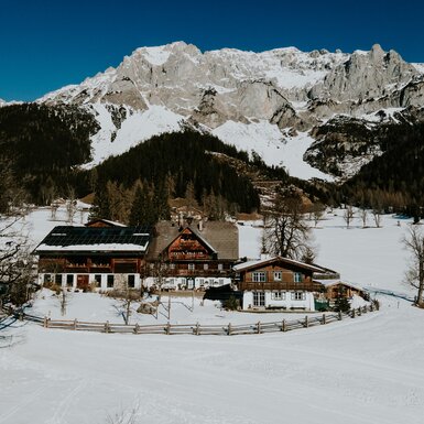 Dorfplatz in einer verschneiten Winterlandschaft mit Bergen im Hintergrund, bestehend aus Holzhäusern mit Schneebedeckten Dächern. | © Urlaub am Bauernhof / Daniel Gollner