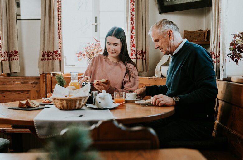 Eine junge Frau und ein älterer Mann sitzen gemeinsam an einem Frühstückstisch in einer gemütlichen Küche, lesen und trinken Kaffee.