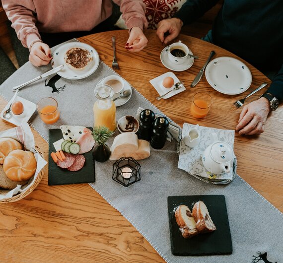 Frühstückstisch mit Kaffee, Saft, Gebäck, Eiern und Nüssen, umgeben von zwei Personen.