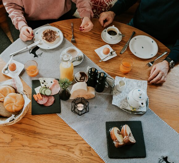 Frühstückstisch mit Kaffee, Saft, Gebäck, Eiern und Nüssen, umgeben von zwei Personen.
