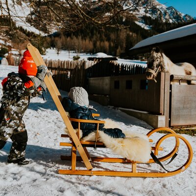 Kind zieht ein Schlitten mit einem Kind, das in einem warmen Mantel und grau gestrickten Mütze sitzt, in einer verschneiten Winterlandschaft.