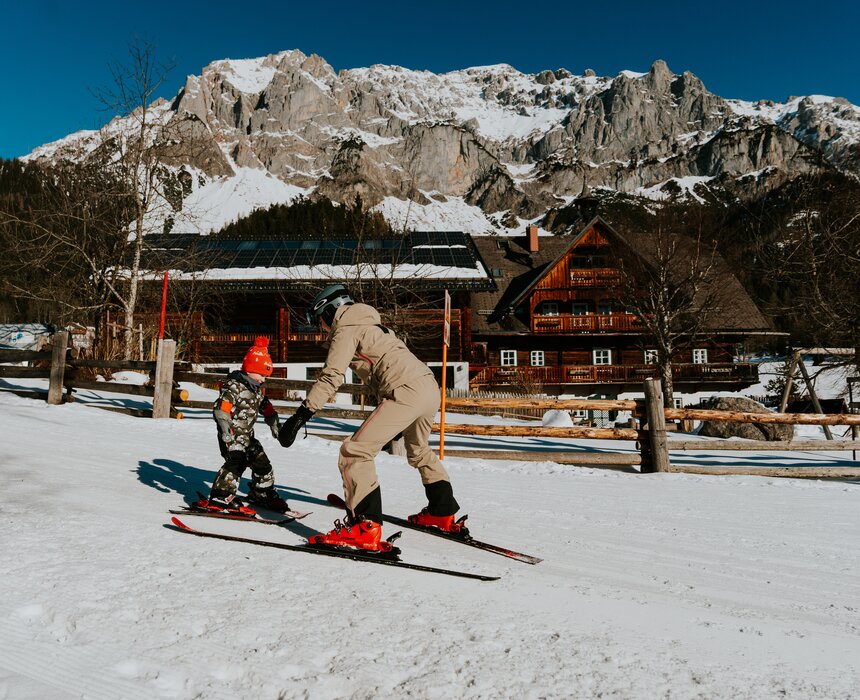 Ein Erwachsener und ein Kind lernen auf Skiern zu stehen, während sie auf einer verschneiten Piste vor einer Holzbank und einer Lodge mit Bergen im Hintergrund trainieren.