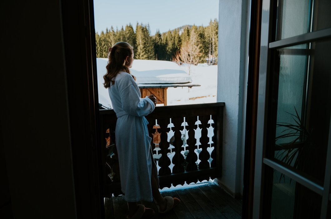 Frau in Bademantel steht am Balkon und blickt auf verschneite Landschaft mit Bäumen.