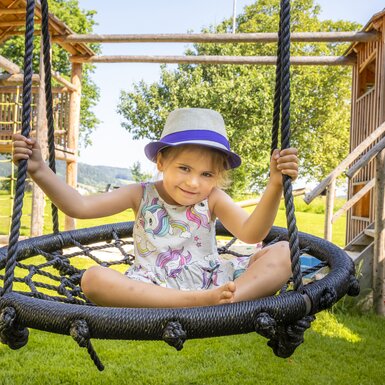 Kleines Mädchen in Sommerkleidung und Hut auf Schaukel in grünem, ländlichen Umfeld mit Holzgebäude im Hintergrund.