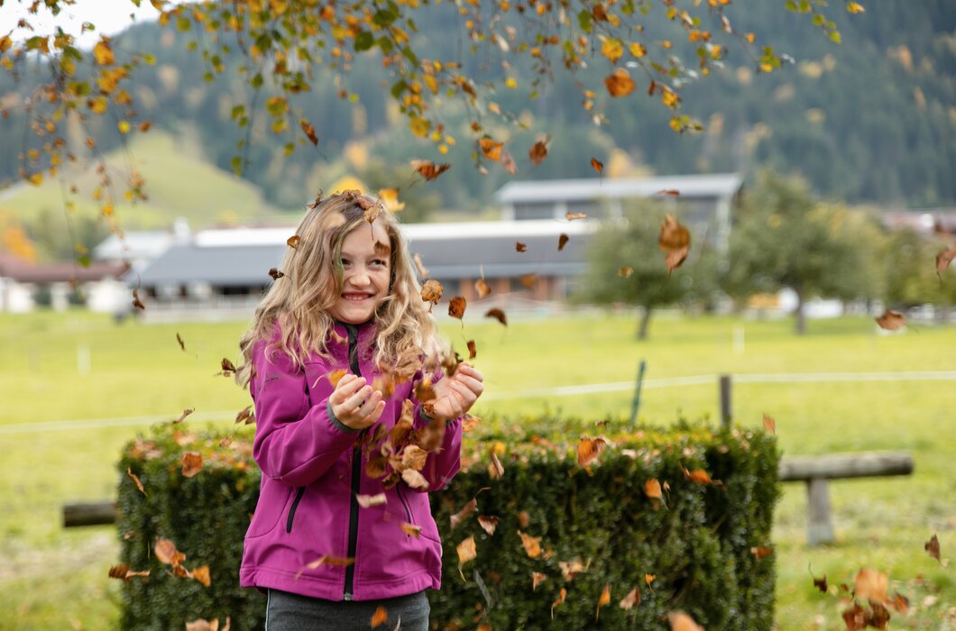 Eine lächelnde junge Frau in lila Jacke, umgeben von herunterfallenden orangefarbenen Blättern in einer parkähnlichen Umgebung mit Gebäuden im Hintergrund. | © Urlaub am Bauernhof / Punkt und Komma