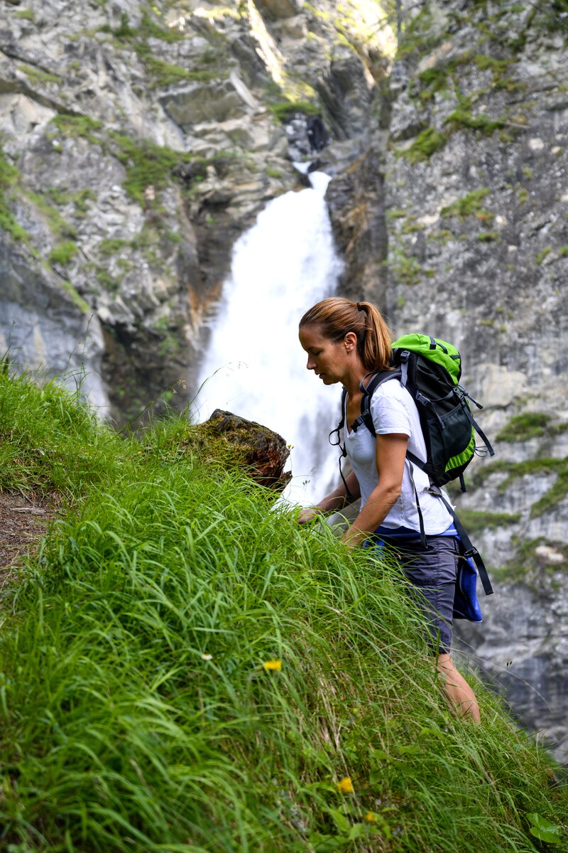 Eine wandernde Person mit grünem Rucksack steht in der Nähe eines hohen Wasserfalls umgeben von üppiger Vegetation. Die Umgebung ist felsig und grün. Die Person scheint den Wasserfall zu betrachten. | © Urlaub am Bauernhof