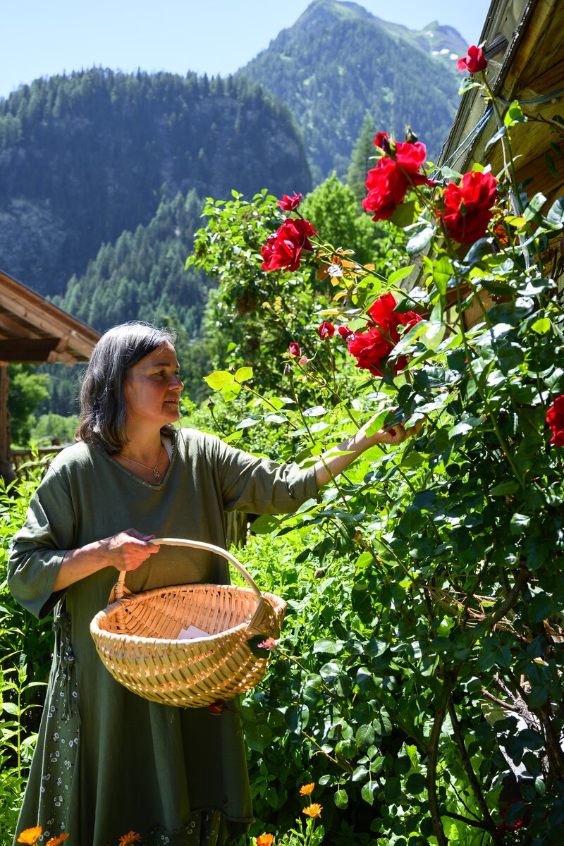 Eine Person in einem grünen Gewand steht in einem üppigen Garten mit roten Rosen und hält einen Korb in der Hand. Im Hintergrund sind bewaldete Berge zu sehen. | © Urlaub am Bauernhof / Punkt und Komma