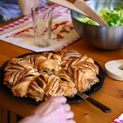 Auf dem Holztisch stehen verschiedene Speisen und Getränke: ein Brotlaib, eine Salatschüssel, ein Glas und ein Backwerk auf einer Platte. Die Platte ist mit einem karierten Tuch bedeckt. | © Urlaub am Bauernhof / Punkt und Komma