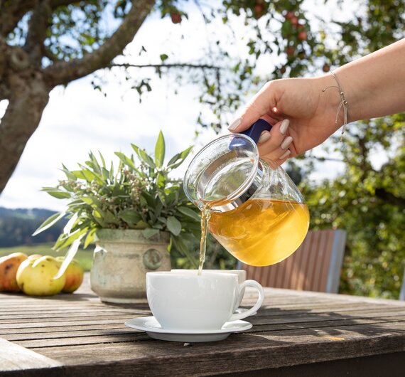 Eine Person gießt Tee in eine Tasse auf einem Holztisch mit Pflanzen und Früchten. Die Umgebung ist von Bäumen umgeben, die eine ländliche und entspannte Atmosphäre schaffen. | © Urlaub am Bauernhof in Österreich / Kimberly Karisch