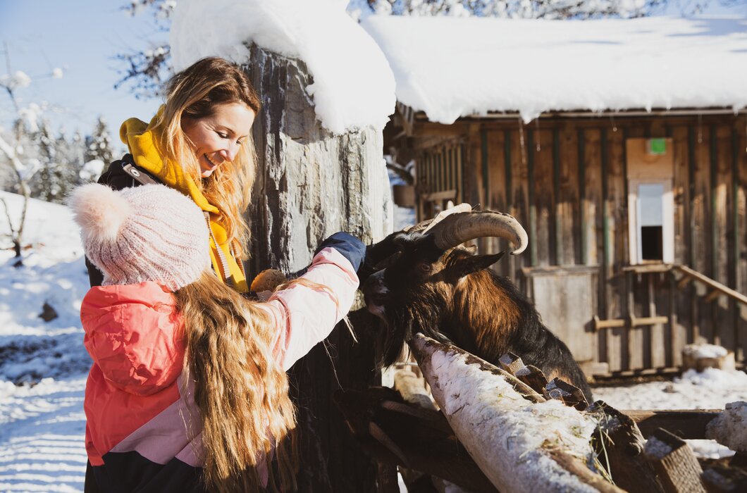 Junge Frau in bunter Winterkleidung streichelt eine Ziege in einer Winterlandschaft mit Holzhäusern im Hintergrund. | © Urlaub am Bauernhof / Pascal Baronit