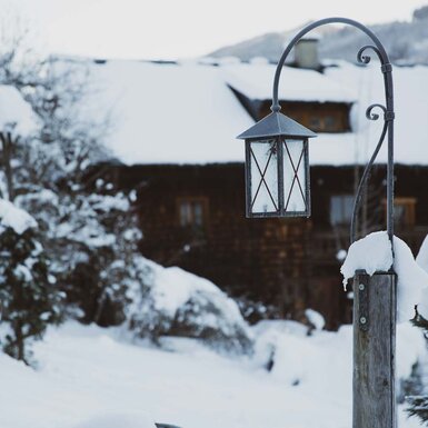 Verschneite Landschaft mit einer alten Laterne vor einem traditionellen Holzhaus. In der Ferne sind weitere verschneite Bäume und Gebäude zu erkennen. | © Urlaub am Bauernhof / Pascal Baronit