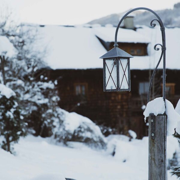 Verschneite Landschaft mit einer alten Laterne vor einem traditionellen Holzhaus. In der Ferne sind weitere verschneite Bäume und Gebäude zu erkennen. | © Urlaub am Bauernhof / Pascal Baronit