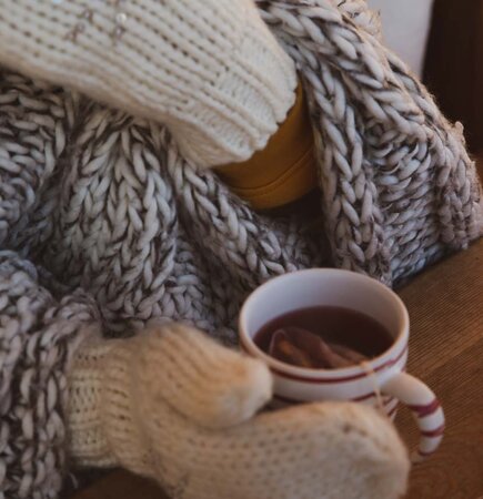 Gestrickter Schal, Handschuhe und eine Tasse dampfenden Kaffees auf einem Holztisch, alles in warmen, mäßigen Farbtönen. | © Urlaub am Bauernhof / Pascal Baronit