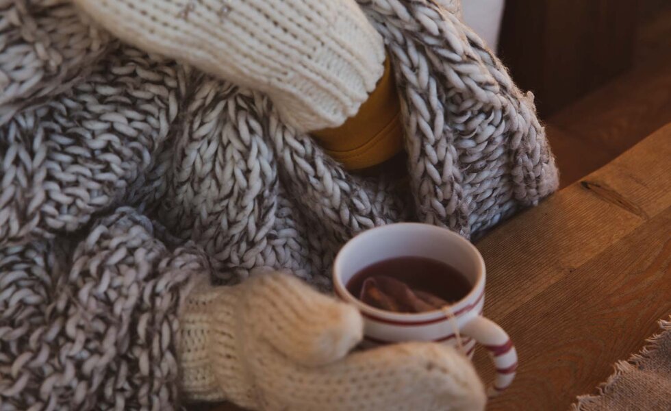 Frau wärmt sich an Tasse Tee | © Urlaub am Bauernhof / Pascal Baronit Gestrickter Schal, Handschuhe und eine Tasse dampfenden Kaffees auf einem Holztisch, alles in warmen, mäßigen Farbtönen. | © Urlaub am Bauernhof / Pascal Baronit