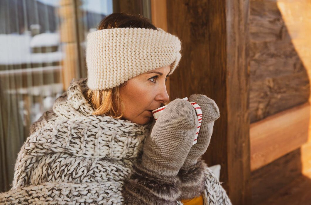 Frau trinkt Tee | © Urlaub am Bauernhof / Pascal Baronit Eine Frau in warmer, gemusterter Kleidung und Mütze trinkt einen heißen Getränk, während sie sich an eine Holzwand lehnt. | © Urlaub am Bauernhof / Pascal Baronit