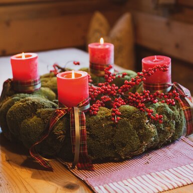 Adventskranz aus Moos mit roten Kerzen, Zierschmuckschleifen und Beeren auf Holztisch. | © Urlaub am Bauernhof / Pascal Baronit