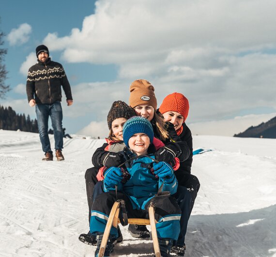 Familie am Schlitten | © Urlaub am Bauernhof / Punkt und Komma Eine Familie genießt einen winterlichen Ausflug: Ein Kind sitzt in einem Schlitten, von Erwachsenen umgeben, die mit warmer Kleidung bekleidet sind. Im Hintergrund eine schneebedeckte Landschaft und Wolken am Himmel. | © Urlaub am Bauernhof / Punkt und Komma