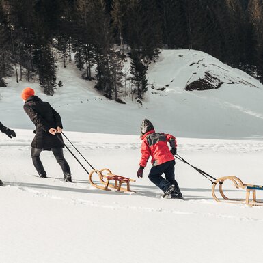 Drei Personen ziehen Schlitten über verschneite Landschaft mit bewaldeten Hügeln im Hintergrund. | © Urlaub am Bauernhof / Punkt und Komma