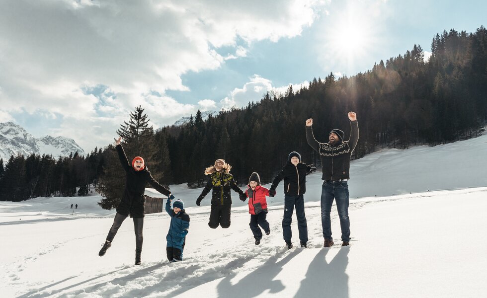 Familie springt im Schnee | © Urlaub am Bauernhof / Punkt und Komma Eine Gruppe von Menschen in Winterkleidung, die freudig mit erhobenen Armen posiert, umgeben von schneebedeckten Bergen und Wäldern unter wolkigem Himmel. | © Urlaub am Bauernhof / Punkt und Komma
