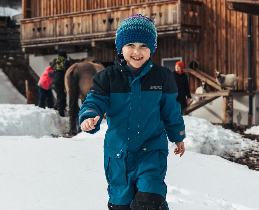Ein lächelndes Kind in einem blauen Winteroutfit spaziert durch den verschneiten Ort, im Hintergrund Holzhäuser und Pferde. | © Urlaub am Bauernhof / Punkt und Komma
