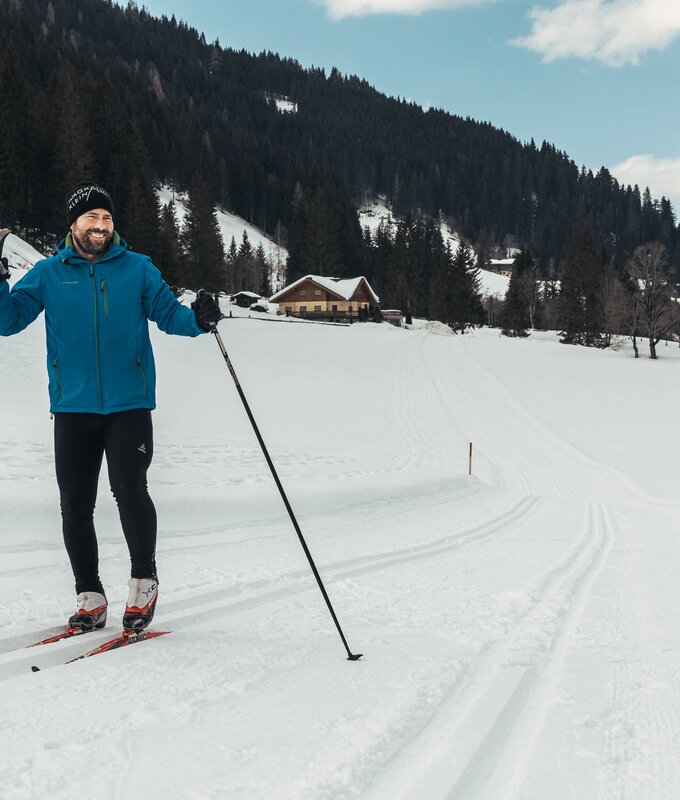 Ein Mann in blauer Jacke und Skiausrüstung auf einer verschneiten Piste mit Berghütte im Hintergrund. | © Urlaub am Bauernhof / Punkt und Komma