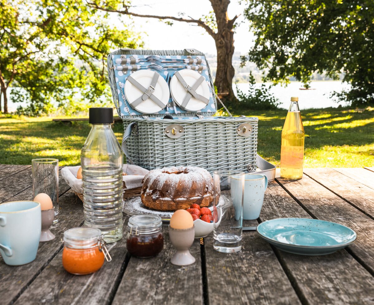 Picknickkorb mit geschirr, kuchen, getränken und früchten auf einem holztisch im garten. schöne sommerliche umgebung mit bäumen und gras als idyllischer hintergrund. | © Urlaub am Bauernhof / Punkt und Komma