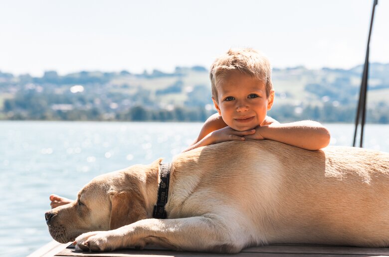 Ein lächelndes Kind, das auf einem Hund liegt und die Umgebung am Ufer betrachtet. Der sonnige Himmel und das ruhige Wasser bilden eine idyllische Szenerie. | © Urlaub am Bauernhof / Punkt und Komma