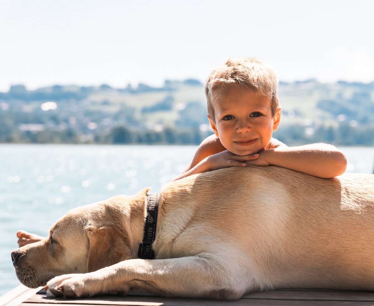 Ein lächelndes Kind, das auf einem Hund liegt und die Umgebung am Ufer betrachtet. Der sonnige Himmel und das ruhige Wasser bilden eine idyllische Szenerie. | © Urlaub am Bauernhof / Punkt und Komma