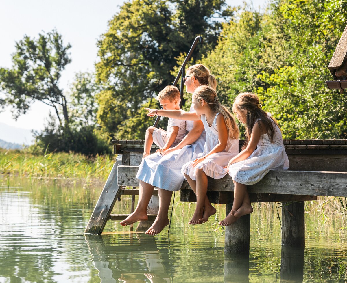 Eine Gruppe von Frauen und Kindern sitzt auf einem Steg über einem See, umgeben von Bäumen und Wiesen. Sie blicken entspannt auf die ruhige Wasserfläche. Das Bild vermittelt eine friedliche, naturverbundene Stimmung. | © Urlaub am Bauernhof / Punkt und Komma