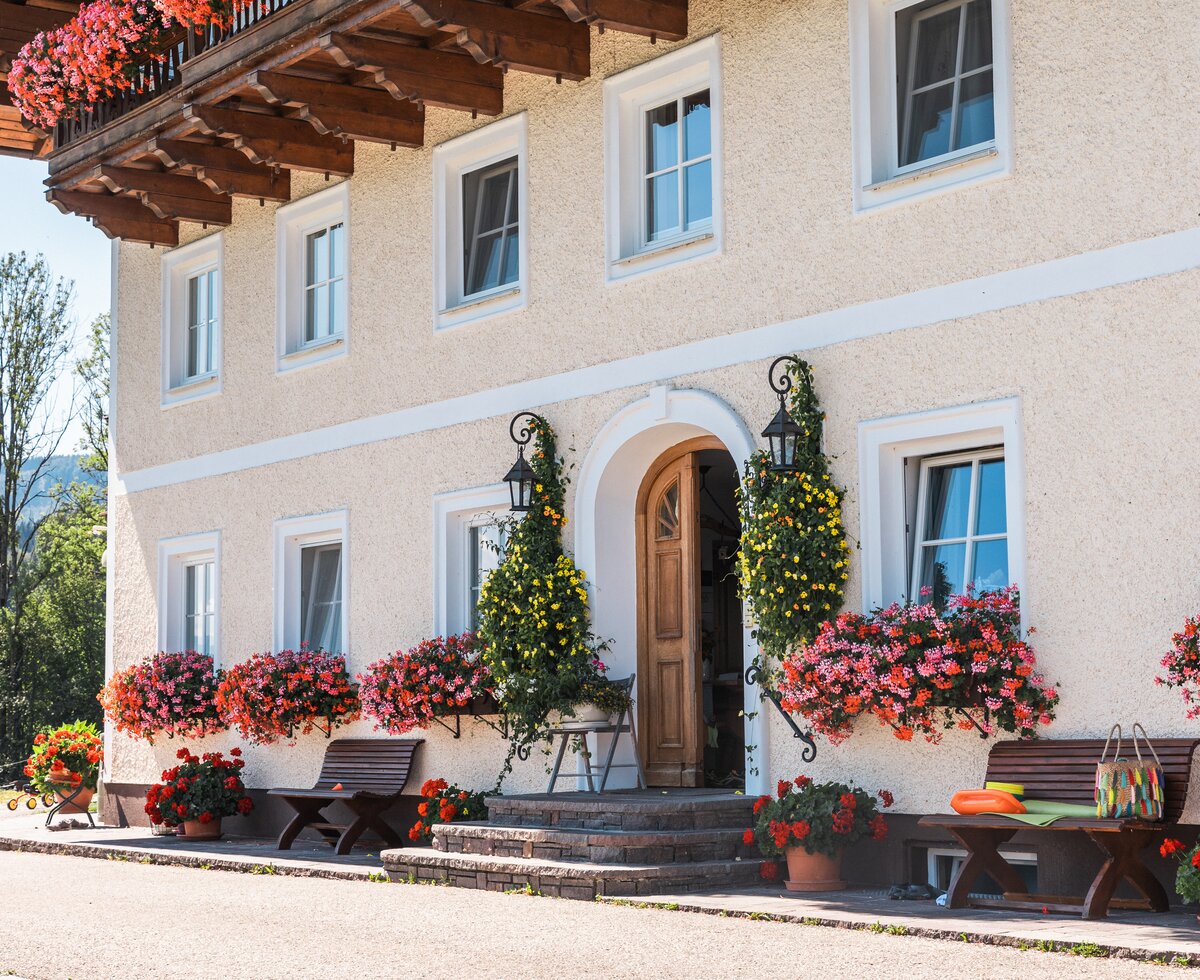 Ein idyllisch restaurierter Bauernhof mit hölzernen Balkongeländern, bunten Blumentöpfen und steinernen Treppen, umgeben von Wiesen und Berglandschaft. | © Urlaub am Bauernhof / Punkt und Komma