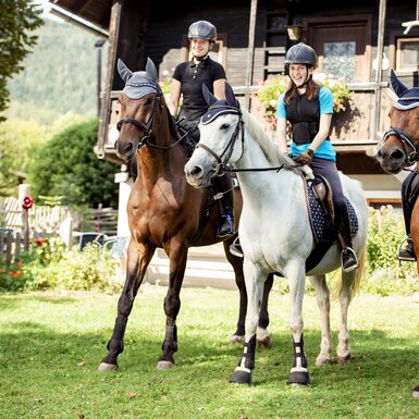 Eine Gruppe von Reitern auf Pferden in einem Garten mit einer Holzhütte im Hintergrund. Die Reiter tragen Helme und Reitkleidung und sitzen auf einem weißen und zwei braunen Pferden. | © Urlaub am Bauernhof Österreich / Andreas Hofer