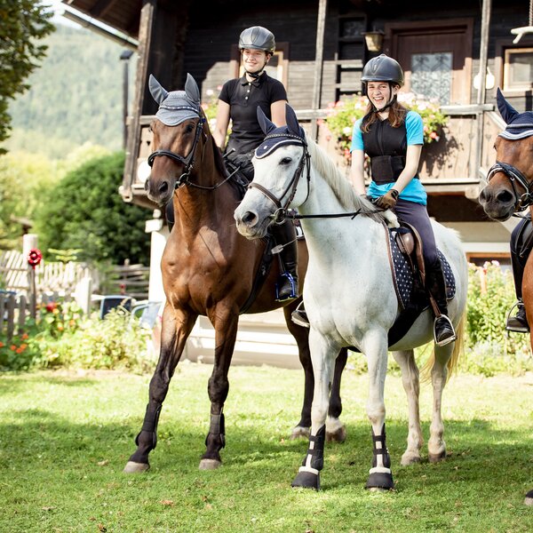 Eine Gruppe von Reitern auf Pferden in einem Garten mit einer Holzhütte im Hintergrund. Die Reiter tragen Helme und Reitkleidung und sitzen auf einem weißen und zwei braunen Pferden. | © Urlaub am Bauernhof Österreich / Andreas Hofer