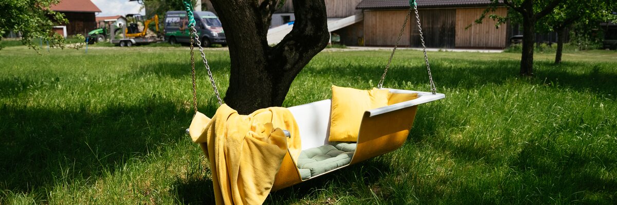 Eine gemütliche, gelbe Hängematte hängt unter einem großen Baum auf einer grünen Wiese. Daneben stehen Autos auf einer Zufahrtsstraße. | © Daniel Gollner / Urlaub am Bauernhof