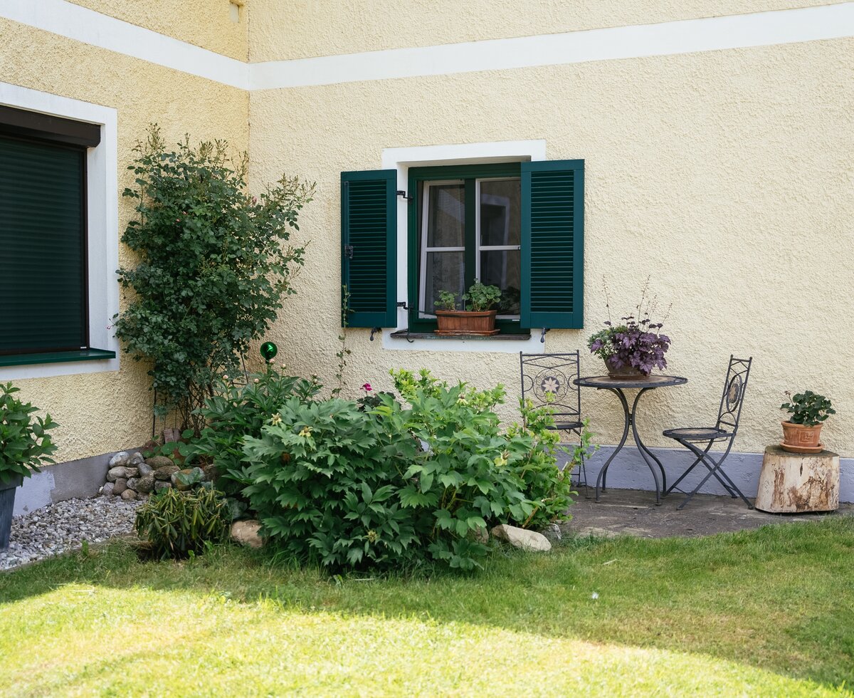 Gemütliches Landhaus mit umlaufenden Grünflächen, geschmückter Fensterbank, Gartentisch und -stuhl sowie Dekorpflanzen auf einer Holzsäule. Insgesamt eine idyllische, natürliche Umgebung. | © Daniel Gollner / Urlaub am Bauernhof