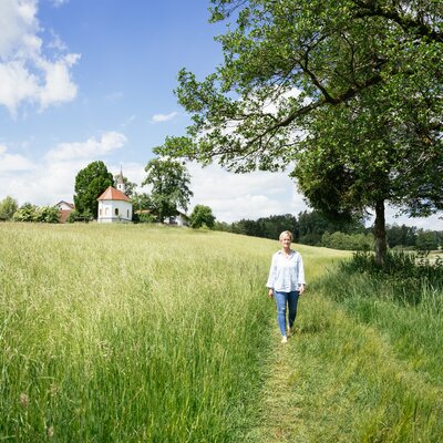 Eine Frau in einer weißen Bluse und Jeans spaziert auf einem Feldweg durch eine grüne, hügelige Landschaft mit Bäumen und einer kleinen Kapelle am Horizont. | © Daniel Gollner / Urlaub am Bauernhof