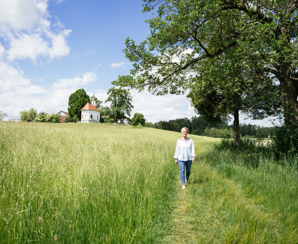 Eine Frau in einer weißen Bluse und Jeans spaziert auf einem Feldweg durch eine grüne, hügelige Landschaft mit Bäumen und einer kleinen Kapelle am Horizont. | © Daniel Gollner / Urlaub am Bauernhof