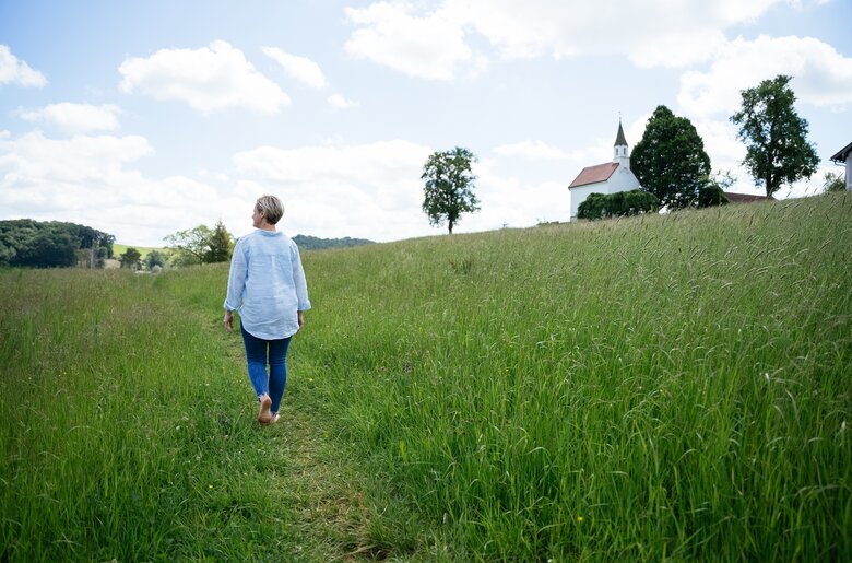 Eine Person in einem blauen Hemd geht über einen grasigen Feldweg, im Hintergrund eine Kirche und Bäume. | © Daniel Gollner / Urlaub am Bauernhof