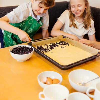 Ein Bub mit grüner Schürze bestreut einen Blechkuchen mit Heidelbeeren und ein Mädchen sieht lächelnd zu, beide sitzen auf der Eckbank in der Küche | © Urlaub am Bauernhof Steiermark / Wolfgang Spekner